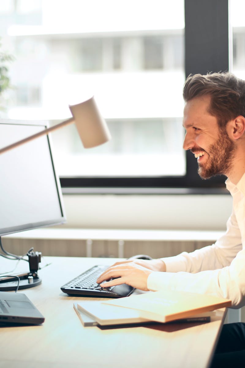 A man smiling while working at an office desk with a computer and natural daylight streaming in through large windows.
